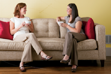 2 ladies sitting on the couch smiling and each holding cup of tea, they are cozy.